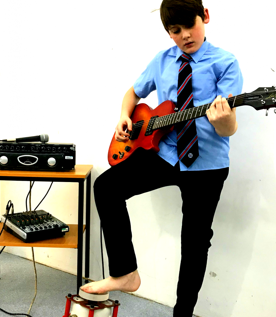 Teenage boy playing a red electric guitar. He has one bare foot placed gently on a vibrotactile foot shaker