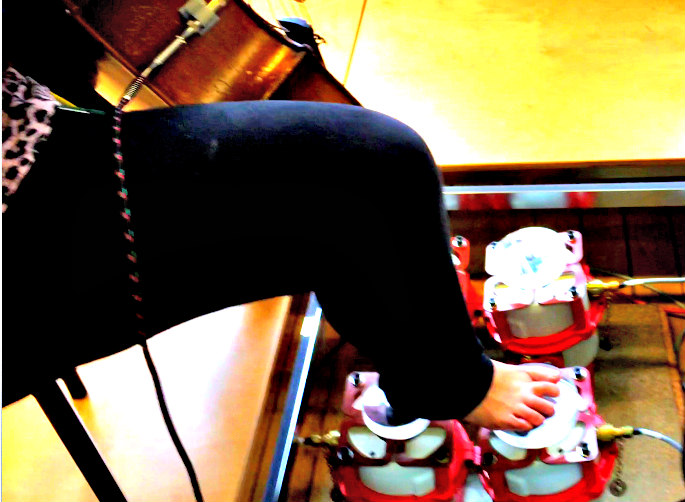 Picture of a woman's leg and part of a cello. The woman's hell and forefoot are resting upon two vibrotactile shakers