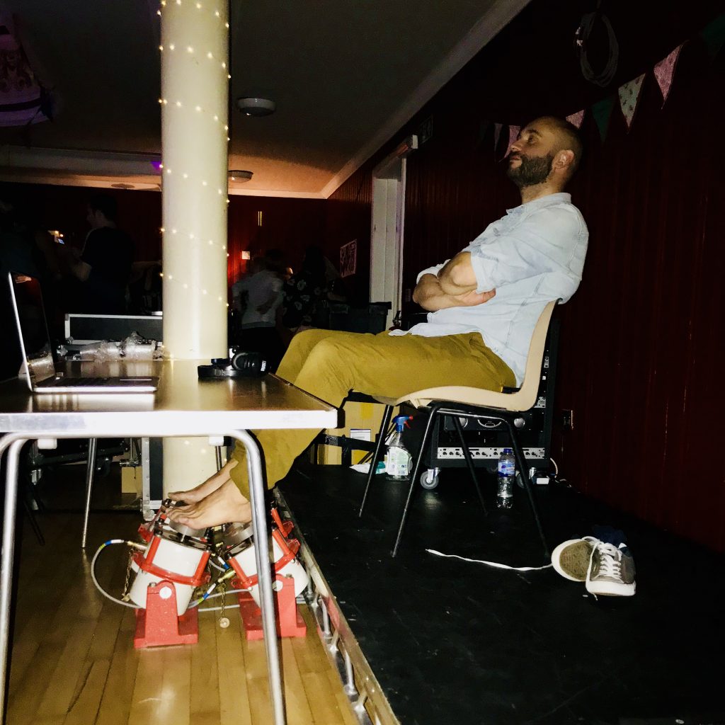 Man seated with bare feet and arms folder, listing to music at a live event. His bare feet are resting on four vibrotactile shakers.