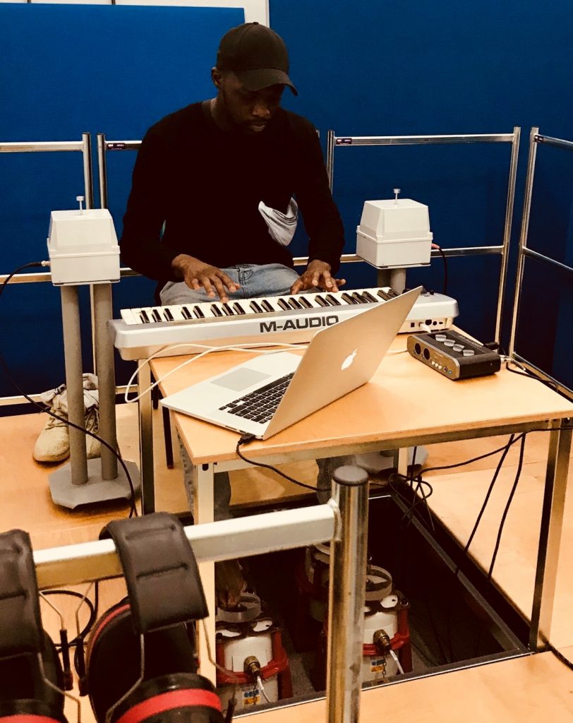 Man seated at a table playing a MIDI keyboard, with bare feet resting on four vibrotactile shakers.