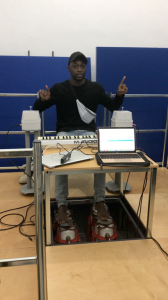 Picture shows man seated at a table. He is signrapping along to a music track. His feet are bare and are placed upon four vibrotactile shakers