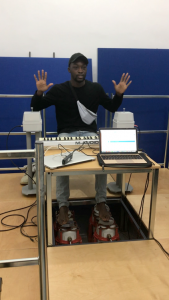 Picture shows man seated at a table. He is signrapping along to a music track. His feet are bare and are placed upon four vibrotactile shakers