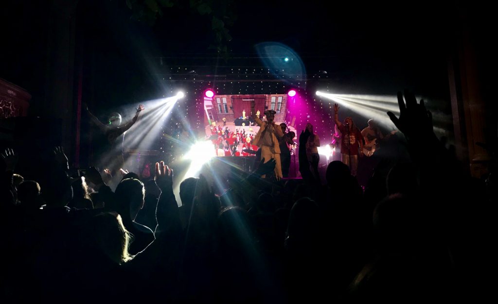 Picture of a band on stage. The lead singer wears a yellow suit and hat. Stage lights are shining out into the audience who have their hands raised.
