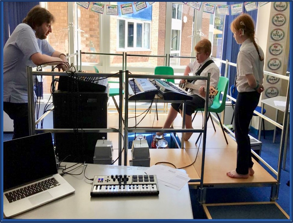 A male teacher watches a young boy around 11 years old playing bass guitar. He is using vibrotactile equipment with his feet so that he can feel the vibrations from the bass guitar. A young girl in school uniform is watching and waiting her turn.. She is wearing a cochlear implant over her left ear.