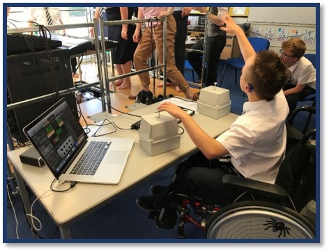 Teenage boy in school uniform in classroom, wearing hearing aid, seated in wheelchair, using vibrotactile equipment with left hand. He is attracting the attention of a teacher using sign language.
