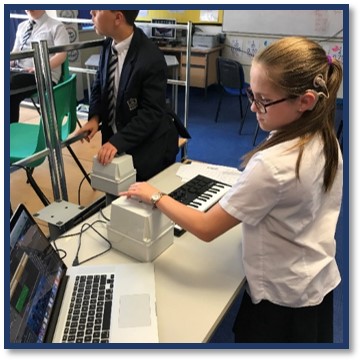 A young girl wearing school uniform in a classroom. She wears a cochlear implant over her left ear. She is using vibrotactile equipment with her left hand and looking at a MacBook screen. Another boy is using vibrotactile equipment with his left hand in the background.