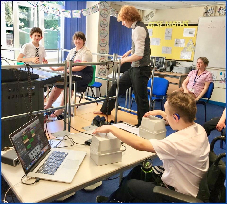 Foreground shows teenage boy in school uniform in classroom, wearing hearing aid, seated in wheelchair, using vibrotactile equipment with both hands. Behind him are two pupils are seated and are using vibrotactile equipment with feet. A male teacher is walking across to them. A female teacher is watching.